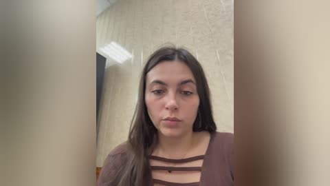 A video of a young woman with long brown hair, wearing a brown top with cut-out straps, standing in a beige-tiled hallway.