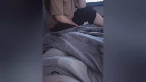 Video of a person in a dimly lit room, sitting on a bed with grey sheets, wearing a brown shirt and black shorts. The person's face is partially out of frame, and the background is dark.