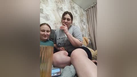 Video of two women in casual attire, sitting on a bed with a plush teddy bear, looking at a laptop screen in a cozy, cluttered room.