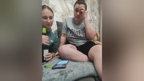 Video of two young women, one holding a green bottle, sitting on a gray blanket. One wears a gray shirt, the other a green one, in a dimly lit room with a patterned wallpaper background.