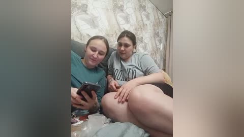 A video shows two overweight women with light skin, one with brown hair in a teal shirt, the other with blonde hair in a gray shirt, sitting on a bed with floral-patterned sheets.