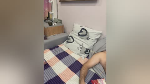 Video of a man lying on a bed with a plaid blanket, gray pillow featuring heart design, and a wooden nightstand with a lamp and books in a small, cozy room.
