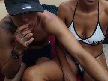 Video of two tan-skinned, shirtless men in bikinis, one with a tattooed hand on his mouth, sitting on a red couch.