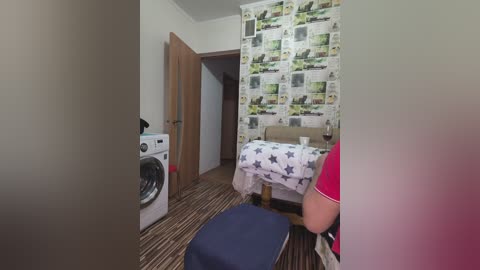 Video of a compact, modern laundry room with a washer, a table with a white star-patterned cloth, and a chair; wallpapered with newspaper clippings.