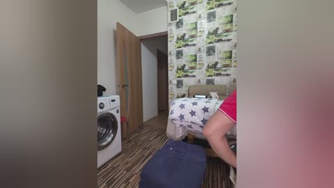 Video of a small, cozy bedroom with a white washing machine, patterned wallpaper, a bed covered in a blue and white star-patterned duvet, and a person in a red shirt.