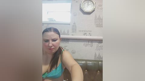 Video of a fair-skinned woman with brown hair, wearing a turquoise lace bra, sitting on a brown leather couch. Background features a white brick wall with London-themed wallpaper and a clock.