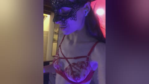 Video of a pale woman with long, straight brown hair wearing a black lace mask and red lace bra, illuminated by purple and red lighting in a dimly lit room.