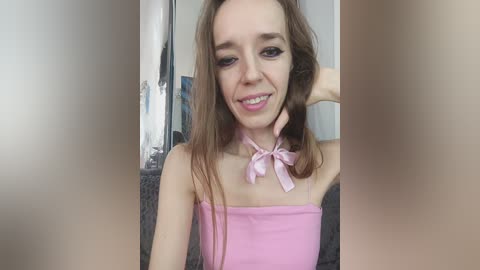 Video of a young woman with fair skin and long brown hair, wearing a pink spaghetti strap top with a bow, smiling and touching her chin.