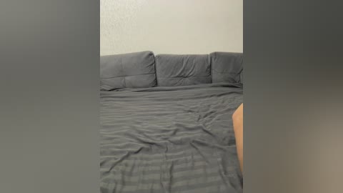 Video of a messy, unmade bed with gray sheets and pillows, against a plain, light-colored wall. The image is framed by blurred vertical edges, suggesting it's taken from a phone camera.