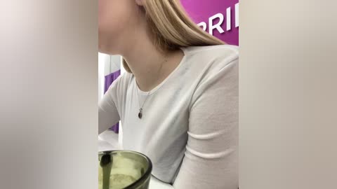 A video of a woman with long blonde hair, wearing a white long-sleeve shirt, standing in a bathroom. She holds a green glass tumbler, with a purple wall and part of a poster with white text visible in the background.