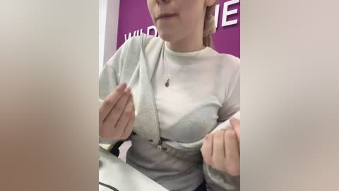 A video of a fair-skinned woman with blonde hair, wearing a white long-sleeved shirt, pulling up the shirt to reveal a light blue bra. The background is a purple wall with partially visible white text.