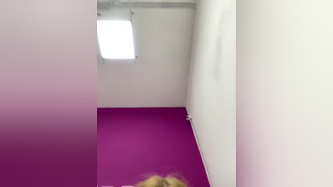 Video of a narrow, white-walled room with a bright, purple floor and a single, rectangular skylight window emitting bright light.