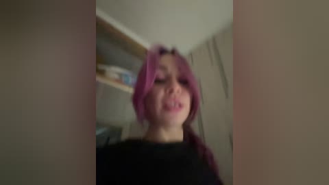A blurry video of a woman with purple hair and a black shirt, partially cropped, standing in a dimly lit room with shelves and a wooden door in the background.