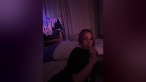 A dimly lit video captures a woman with light skin and blonde hair, wearing a black shirt, lying on a bed. She's holding a phone to her mouth, looking toward the camera. The background includes a partially visible wooden chair and a window with sheer curtains.