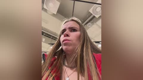 A video of a blonde woman with long, straight hair, wearing a red top and silver necklace, taken from a low angle in a dimly lit, cluttered room with visible ceiling fans and hanging clothes.