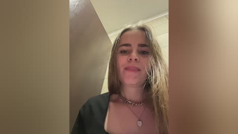 Video of a young woman with long, wet, blonde hair, wearing a black top and necklace, standing in a narrow corridor with beige walls, looking directly at the camera.