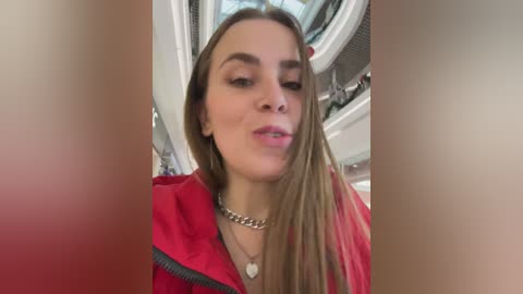 Video of a young woman with long, straight brown hair, wearing a red jacket, smiling, inside a car with a white interior.