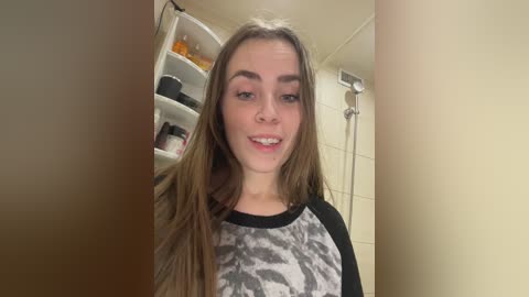 A video of a young Caucasian woman with long, straight brown hair, wearing a black and white graphic tee, smiling in a bathroom with a shower and shelves filled with toiletries.