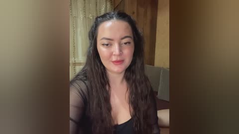 Video of a smiling, fair-skinned woman with long, wavy brown hair, wearing a sheer black top, in a cozy room with wooden walls and a window with lace curtains.