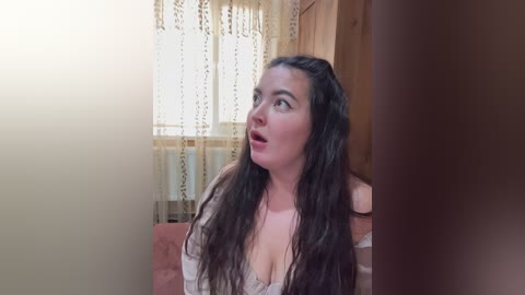 A video of a woman with long, wavy dark hair, wearing a white off-shoulder top, seated indoors near a window with lace curtains. Her expression is surprised or shocked, with wide eyes and an open mouth.