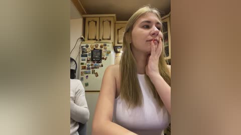 Video of a blonde woman with medium build, wearing a white tank top, touching her chin, standing in a beige kitchen with wooden cabinets and a refrigerator adorned with magnets.