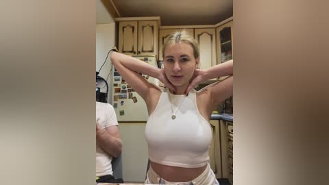 A video of a blonde woman with fair skin, medium breasts, and medium build, wearing a white crop top and shorts, posing with hands behind head in a cozy, wooden kitchen with cabinets and a fridge.