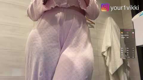 Video of a woman in a light purple satin robe, standing in a modern bathroom with beige tiled walls and a hanging towel.