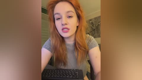 Video of a fair-skinned, red-haired woman wearing a gray t-shirt, typing on a black keyboard in a room with beige walls and floral wallpaper.
