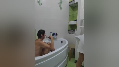 Video of a nude man with short brown hair sitting in a white bathtub, legs crossed, holding a bottle, in a minimalist, white-tiled bathroom with green accents.