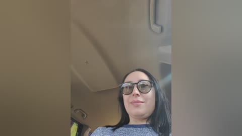 Video of a smiling woman with straight black hair, wearing black-rimmed glasses, gray sweater, and a yellow backpack, standing in a beige room with a vent on the ceiling.