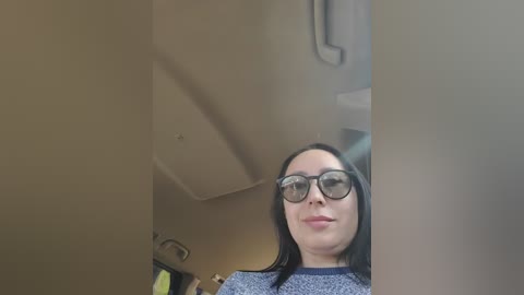 A video of a woman with long black hair, wearing large black-rimmed glasses and a grey sweater, taken from a low angle in a dimly lit room.