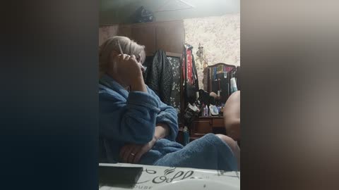A video of an elderly woman with gray hair in a blue robe, seated in a cluttered, dimly lit bedroom with floral wallpaper and clothes hanging in the background.