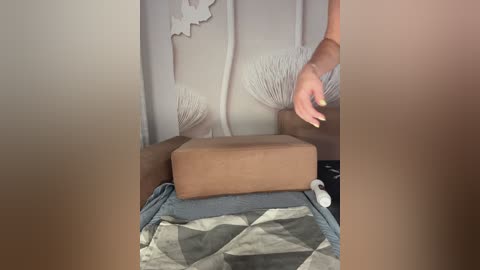 A video of a person's arm reaching towards a wooden box on a bed with a patterned quilt. The background shows a white wall with abstract art.