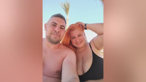 Video of a shirtless man with short hair and a woman with red hair, both smiling, wearing black tank tops, standing outdoors with a clear blue sky and palm trees in the background.