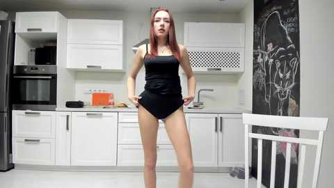 Video of a slender, red-haired woman in a black tank top and shorts standing in a modern, white kitchen with black graffiti art on the wall.
