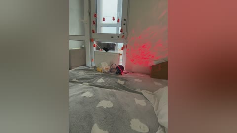 A video of a small, dimly lit room with a bed covered in gray and white patterned sheets. The walls are white, and a window with red light streaming in is visible. Toys and a backpack are scattered on the floor.