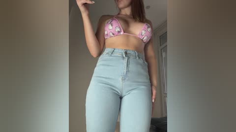 Video of a light-skinned woman with brown hair, wearing a pink bikini with Minnie Mouse pattern and high-waisted light blue jeans, indoors with beige walls and a door in the background.