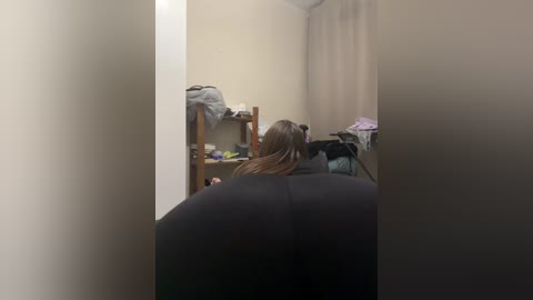 Video of a woman with long brown hair, seated in a dark salon chair, facing away from the camera. The background features beige walls, a wooden shelf with clothes, and a curtain.