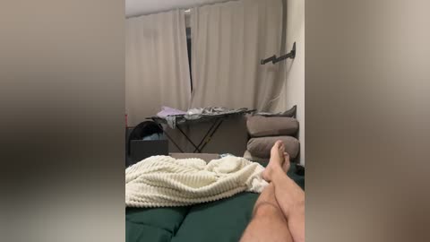 Video of a bedroom with beige curtains, a green bed, a white knitted blanket, and a man's bare feet on the bed.