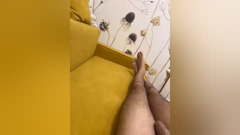 A video of a man's bare legs and feet extending from a mustard-yellow sofa, against a wall adorned with black-and-white floral wallpaper.