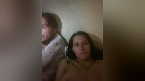 A video shows a couple lying in bed, blurred by a vertical object. The man has a beard, and the woman has dark hair, tattoos, and a necklace.