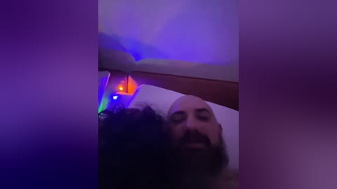 A video of a man with a full beard, partially obscured by a colorful, abstract light display on a wall. The man's face is partially illuminated, creating a dramatic and moody atmosphere.