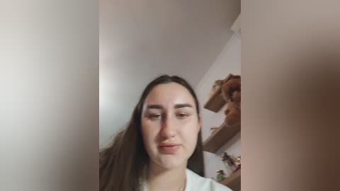 Video of a young woman with long, straight brown hair and light skin, wearing a white top, looking into a mirror in a simple, white-walled room.