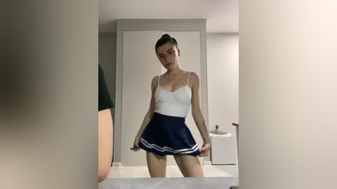 A video of a slender, light-skinned woman with a high ponytail, wearing a white lace camisole and a navy blue pleated skirt, standing in a modern, minimalist bathroom.