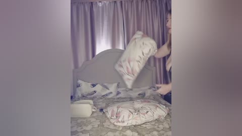 Video of a woman in a beige tank top standing on a bed with a floral-patterned duvet, fluffing pillows. Pink curtains in the background.