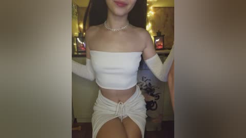 Video of a young woman with light skin and long dark hair, wearing a white off-the-shoulder crop top and matching high-waisted shorts, posing indoors with string lights and lanterns in the background.