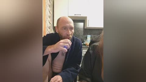 Video of a bald, bearded man in a dark robe, drinking from a blue water bottle, seated on a couch in a modern kitchen with white cabinets, granite countertops, and a brick wall.