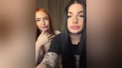 Video of two women with fair skin and long, straight hair: one with red hair, topless, and the other with black hair, wearing a black T-shirt, both puckering their lips in a playful manner.