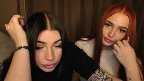 Video of two women, one with long black hair, one with long red hair, both smiling, touching their cheeks, in a dimly lit room with brown curtains.