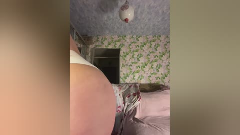 Video of a woman's back, featuring light skin, lying on a bed with pink sheets and floral-patterned wallpaper. The ceiling is textured and adorned with a white, spherical light fixture.